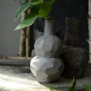 K&ouml;nnte beinhalten: Eine graue Keramikvase mit strukturierter Oberfl&auml;che. Die Vase hat einen runden K&ouml;rper und einen schmalen Hals. Sie steht auf einer Holzoberfl&auml;che mit gr&uuml;nen Bl&auml;ttern.