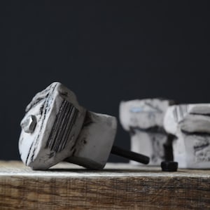 Może przedstawiać: Zbliżenie dwóch szarych i białych gałek do szafek o teksturowanym, skalistym wyglądzie. Każda gałka ma czarny trzpień i srebrny akcent. Gałki znajdują się na drewnianej powierzchni na ciemnym tle.