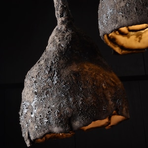 May include: Close-up of three textured, dark brown pendant lights with a rough, organic appearance. The interior of the lampshades glows with a warm, golden light, creating a striking contrast against the dark exterior. The lamps are suspended from the ceiling.
