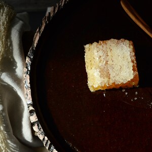 以下が含まれることがあります： 濃い茶色のセラミックプレートの上に置かれたハチミツの巣。ハチミツの巣は薄い茶色で、白くワックスのような質感です。