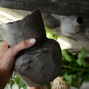 K&ouml;nnte beinhalten: Eine dunkelgraue Keramikvase mit abgebrochenem Rand, die in einer Hand gehalten wird. Die Vase hat eine raue, strukturierte Oberfl&auml;che und ist leicht deformiert.