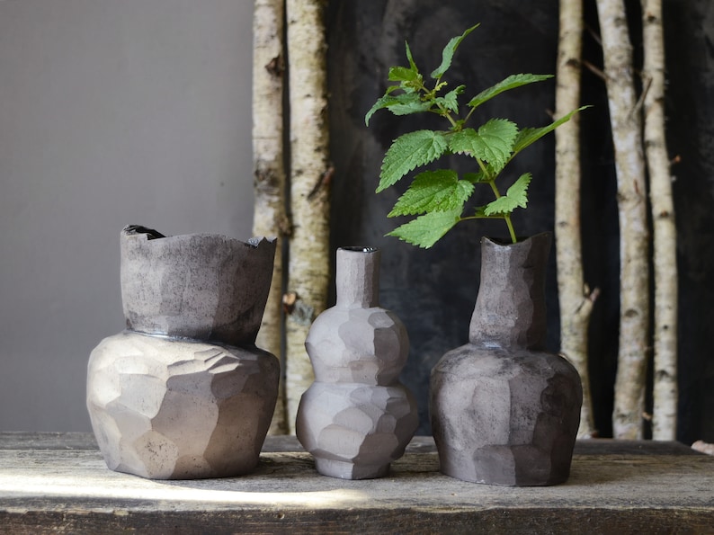 K&ouml;nnte beinhalten: Drei dunkelgraue Keramikvasen mit rauen, strukturierten Oberfl&auml;chen. Die Vase in der Mitte hat eine runde, bauchige Form. Die Vase rechts hat einen schmalen Hals und einen breiteren Boden. Die Vase links hat eine breite &Ouml;ffnung und einen abgerundeten Boden. Ein Zweig mit gr&uuml;nen Bl&auml;ttern befindet sich in der Vase rechts.