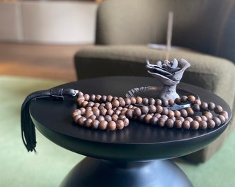 May include: A dark brown wooden mala bead necklace with a black tassel sits on a black table next to a gray ceramic rose incense holder.