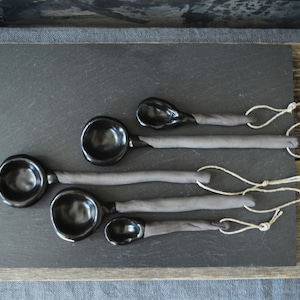 May include: Set of five black ceramic spoons with gray handles and twine ties. The spoons are arranged in a graduated size order on a black slate surface.