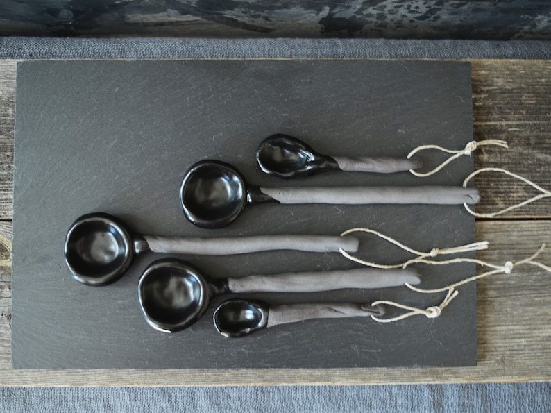 May include: Set of five black ceramic spoons with gray handles and twine ties. The spoons are arranged in a graduated size order on a black slate surface.