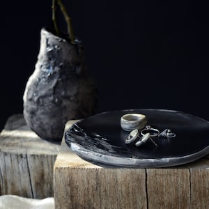 Op de afbeelding: Een zwart keramiek bord met een zilveren ring en oorbellen op een houten oppervlak. Het bord heeft een getextureerd oppervlak en een licht verhoogde rand. De ring en oorbellen zijn eenvoudig en elegant.