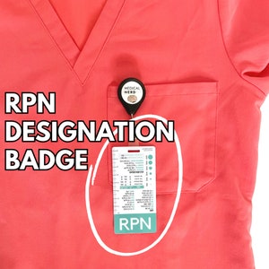Peut inclure: Un haut de blouse rose avec une poche sur le côté gauche. Un badge avec le texte "RPN" est attaché à la poche avec un porte-badge rétractable. Le porte-badge a un petit charme rond avec le texte "MEDICAL NERD".