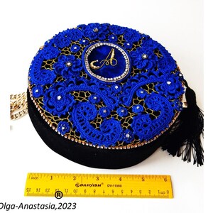 K&ouml;nnte beinhalten: Eine runde blaue und goldene Clutch mit einem schwarzen Quastenanh&auml;nger. Die Tasche ist mit filigraner Spitze und Strasssteinen verziert. Die Tasche hat einen goldenen Kettenriemen und ein blaues Samtst&uuml;ck mit einem goldenen Buchstaben "A" in der Mitte.