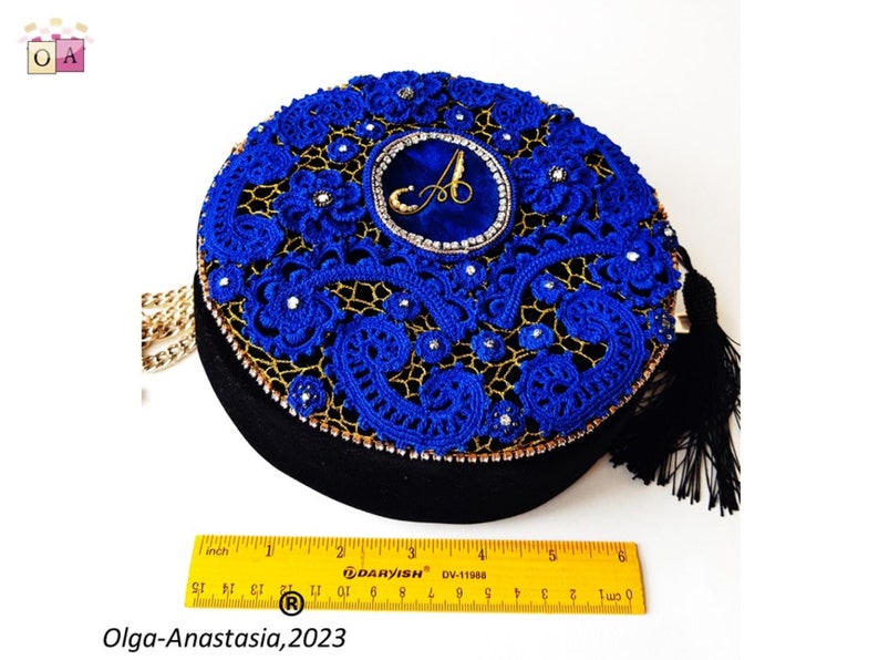 K&ouml;nnte beinhalten: Eine runde blaue und goldene Clutch mit einem schwarzen Quastenanh&auml;nger. Die Tasche ist mit filigraner Spitze und Strasssteinen verziert. Die Tasche hat einen goldenen Kettenriemen und ein blaues Samtst&uuml;ck mit einem goldenen Buchstaben "A" in der Mitte.