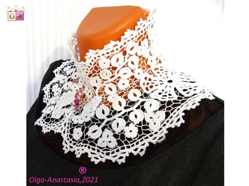 Puede incluir: Collar de encaje blanco tejido a crochet con un delicado patr&oacute;n floral. El collar se lleva sobre una camisa negra y tiene un peque&ntilde;o broche de flor rosa.