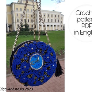 K&ouml;nnte beinhalten: Eine blaue runde H&auml;kel-Tasche mit einer goldenen Kettenkette und einem schwarzen Quasten. Die Tasche hat ein blaues Samtzentrum mit einem goldenen Buchstaben "A" darauf gestickt. Die Tasche befindet sich vor einem Geb&auml;ude mit gr&uuml;nem Gras.