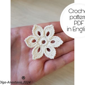 Peut inclure: Une fleur au crochet de couleur crème, avec six pétales et un anneau central de points. L'image comprend le texte "Crochet pattern PDF in English". La fleur est tenue dans une main.