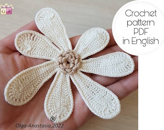 May include: A crocheted flower with six petals in a cream color. The flower is held in a hand. The text "Crochet pattern PDF in English" is in a white circle on the right side of the image.