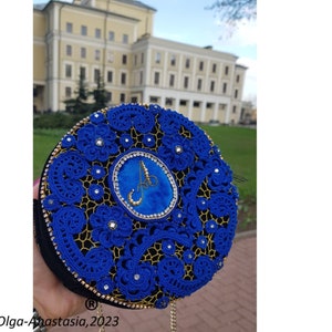 K&ouml;nnte beinhalten: Eine runde blaue und goldene Clutch mit einem Perlenmuster und einem gro&szlig;en blauen Edelstein in der Mitte. Die Clutch hat einen goldenen Kettenriemen und der Buchstabe "A" ist in Gold auf den Edelstein gestickt.