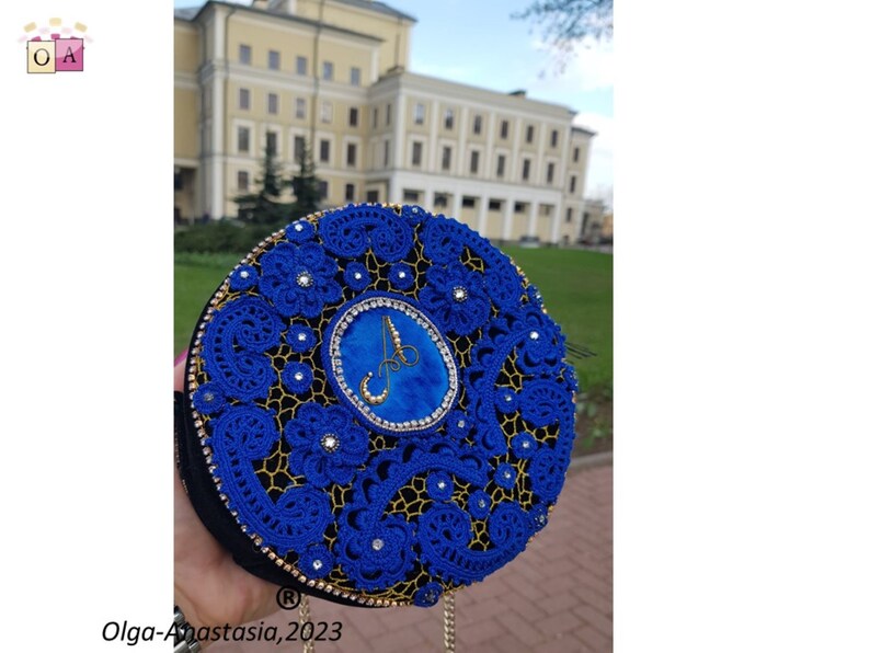 K&ouml;nnte beinhalten: Eine runde blaue und goldene Clutch mit einem Perlenmuster und einem gro&szlig;en blauen Edelstein in der Mitte. Die Clutch hat einen goldenen Kettenriemen und der Buchstabe "A" ist in Gold auf den Edelstein gestickt.