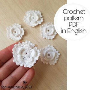 May include: Five white crocheted flowers, each with a small center hole. The flowers are arranged on a light brown wooden surface. The text "Crochet pattern PDF in English" is written in black on a white circle.