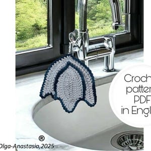 Op de afbeelding: Een blauwe en grijze gehaakte vaatdoek die aan een chromen keukenkraan hangt. De vaatdoek is bladvormig. De tekst "Crochet pattern PDF in English" is zichtbaar in de afbeelding.