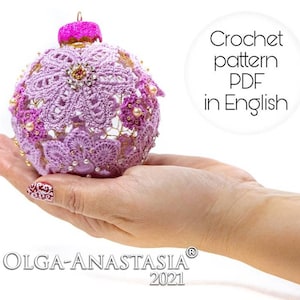 Può includere: Una mano tiene in mano un ornamento natalizio di colore viola lavorato a uncinetto con decorazioni rosa e dorate. L'ornamento è decorato con perline bianche e fiori rosa. Il testo "Crochet pattern PDF in English" è visibile nell'immagine.