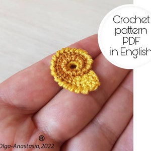 May include: A crocheted yellow and orange nautilus shell, a small crocheted sea creature, in a person's hand. Crochet pattern PDF in English.