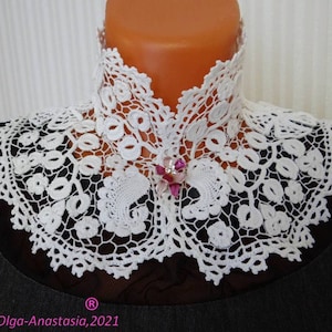Puede incluir: Collar de encaje blanco tejido a crochet con un delicado patr&oacute;n floral. El collar tiene un escote alto y est&aacute; adornado con un broche de flor rosa.
