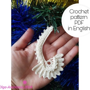 Puede incluir: Adorno de ganchillo color crema con forma de concha estilizada. El adorno se sostiene en una mano sobre un fondo de oropel azul y dorado. El texto de la imagen dice "Crochet pattern PDF in English."