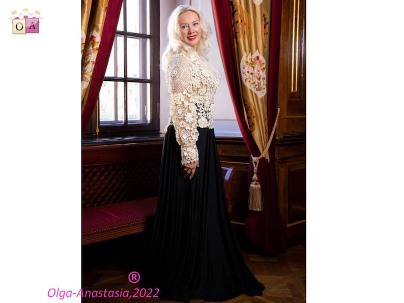 May include: A woman wearing a white crocheted top with a floral design and a black maxi skirt. She is standing in front of a window with red curtains and a wooden door.