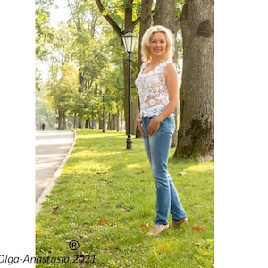 May include: A woman in a white crocheted top and blue jeans stands in a park with trees and a lamppost in the background.