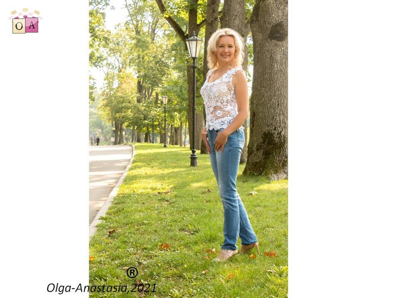 May include: A woman in a white crocheted top and blue jeans stands in a park with trees and a lamppost in the background.