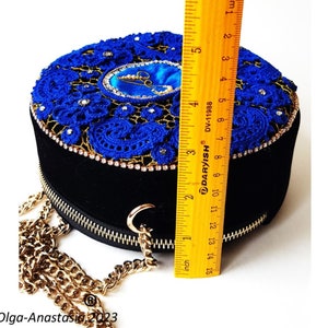 K&ouml;nnte beinhalten: Eine runde Handtasche aus schwarzem Samt mit einem blauen H&auml;kel-Oberteil und einem goldenen Kettenriemen. Das Oberteil ist mit einem blauen und goldenen Design mit Strasssteinen verziert. Die Tasche hat einen Durchmesser von etwa 15 cm.