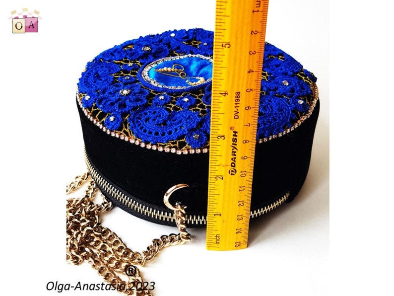 K&ouml;nnte beinhalten: Eine runde Handtasche aus schwarzem Samt mit einem blauen H&auml;kel-Oberteil und einem goldenen Kettenriemen. Das Oberteil ist mit einem blauen und goldenen Design mit Strasssteinen verziert. Die Tasche hat einen Durchmesser von etwa 15 cm.