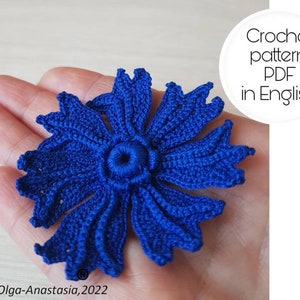 Può includere: Un fiore blu all'uncinetto con un cerchio centrale. Il fiore è tenuto in una mano. Il testo "Crochet pattern PDF in English" è visibile nell'immagine.