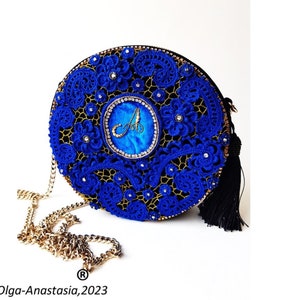 K&ouml;nnte beinhalten: Eine runde blaue und goldene Abendtasche mit einem schwarzen Quastenanh&auml;nger. Die Tasche ist mit filigraner Spitze und Strasssteinen verziert. Die Tasche hat einen goldenen Kettenriemen und ein blaues und goldenes rundes Emblem mit dem Buchstaben "A" in Gold.