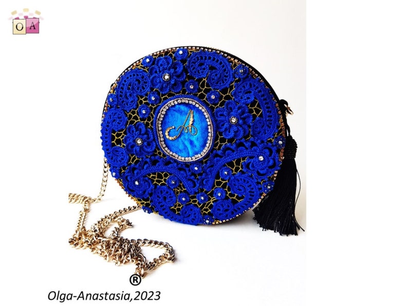 K&ouml;nnte beinhalten: Eine runde blaue und goldene Abendtasche mit einem schwarzen Quastenanh&auml;nger. Die Tasche ist mit filigraner Spitze und Strasssteinen verziert. Die Tasche hat einen goldenen Kettenriemen und ein blaues und goldenes rundes Emblem mit dem Buchstaben "A" in Gold.