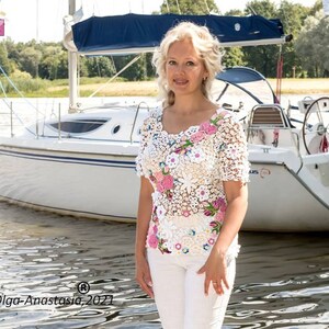 Puede incluir: Una mujer lleva una blusa de crochet blanca con flores rosas y blancas. La blusa tiene mangas cortas y un escote redondo. La mujer est&aacute; de pie frente a un velero blanco.