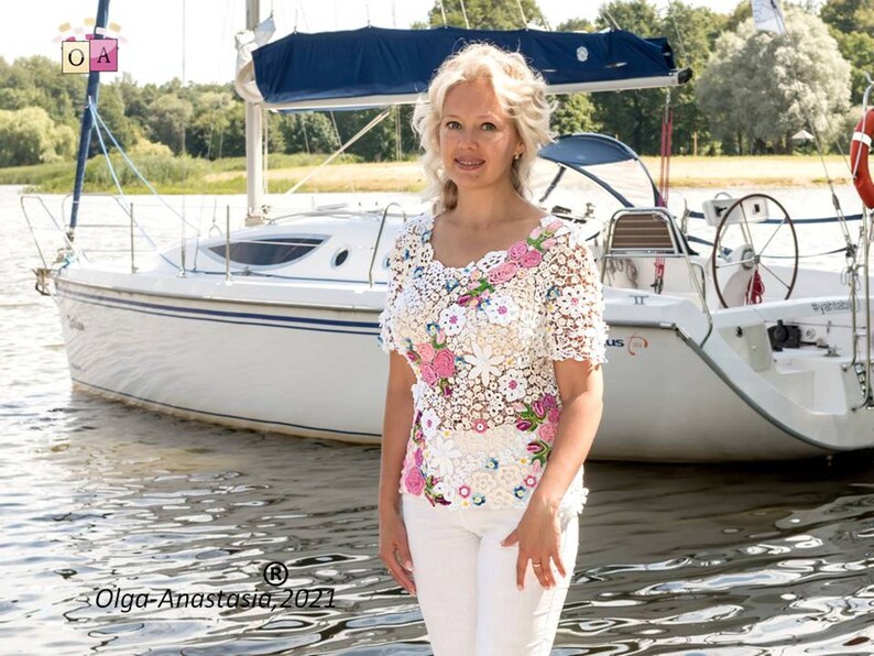 Puede incluir: Una mujer lleva una blusa de crochet blanca con flores rosas y blancas. La blusa tiene mangas cortas y un escote redondo. La mujer est&aacute; de pie frente a un velero blanco.