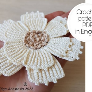 May include: A crocheted white flower with a brown center. The flower is made with a single crochet stitch and has a layered design. The image includes the text "Crochet pattern PDF in English" and "Olga-Anastasia, 2022".