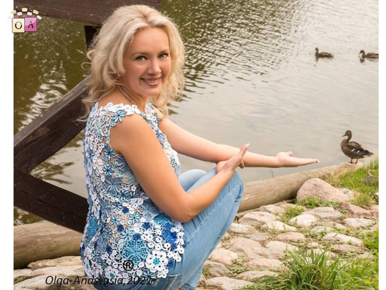 May include: A woman wearing a blue and white crocheted top with a floral design, sitting on a wooden bridge by a pond with ducks. The text "Olga-Anastasia, 2021" is visible in the bottom left corner of the image.