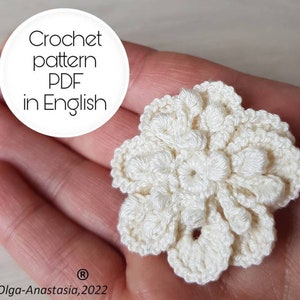 Könnte beinhalten: Nahaufnahme einer cremefarbenen, gehäkelten Blume, die in einer Hand gehalten wird. Die Blume hat ein detailliertes, strukturiertes Design mit mehreren Blütenblättern. Der Text auf dem Bild lautet "Crochet pattern PDF in English".