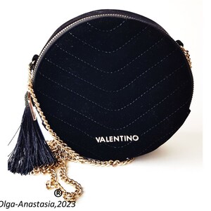 K&ouml;nnte beinhalten: Eine schwarze runde Umh&auml;ngetasche aus Samt mit einer goldenen Kettenkette und einem schwarzen Quasten. Die Tasche hat ein gestepptes Design und das Wort "VALENTINO" ist auf der Vorderseite aufgedruckt.