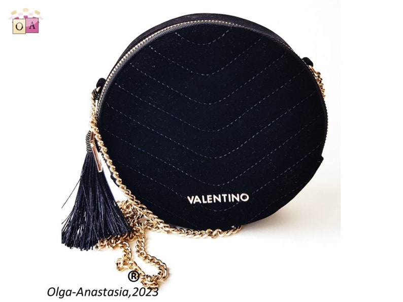 K&ouml;nnte beinhalten: Eine schwarze runde Umh&auml;ngetasche aus Samt mit einer goldenen Kettenkette und einem schwarzen Quasten. Die Tasche hat ein gestepptes Design und das Wort "VALENTINO" ist auf der Vorderseite aufgedruckt.
