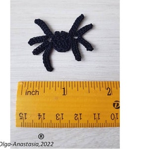 Könnte beinhalten: Eine kleine, schwarze gehäkelte Spinne mit acht Beinen, neben einem Lineal platziert. Das Lineal zeigt Messungen in Zoll an. Die Spinne ist etwa 2,5 cm lang. Das Bild enthält den Text "Olga-Anastasia, 2022".