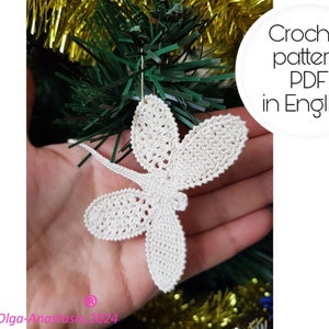 Könnte beinhalten: Handgefertigter, cremefarbener Häkel-Libellen-Ornament. Die Libelle hat detaillierte Flügel und einen Körper. Das Bild enthält den Text "Crochet pattern PDF in English". Der Schmuck hängt vor einem Weihnachtsbaum.