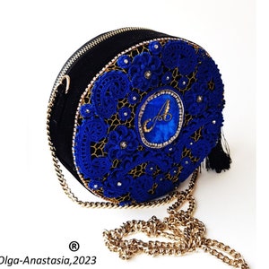K&ouml;nnte beinhalten: Eine runde schwarze Handtasche mit einem blauen H&auml;kelmuster und einem goldenen Kettenriemen. Die Tasche hat einen goldenen Quastenanh&auml;nger und einen in Blau und Gold gestickten Buchstaben "A" in der Mitte.