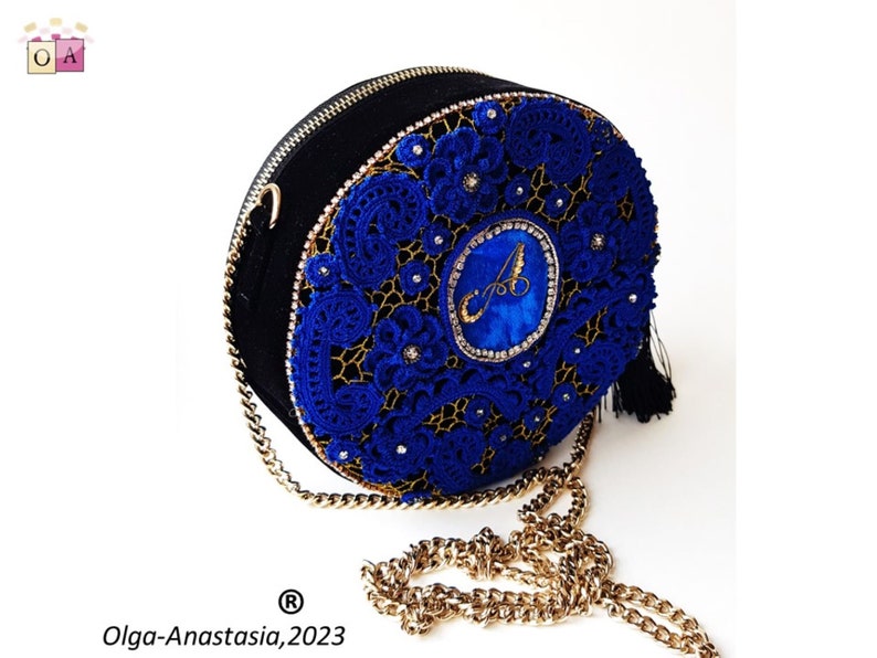 K&ouml;nnte beinhalten: Eine runde schwarze Handtasche mit einem blauen H&auml;kelmuster und einem goldenen Kettenriemen. Die Tasche hat einen goldenen Quastenanh&auml;nger und einen in Blau und Gold gestickten Buchstaben "A" in der Mitte.