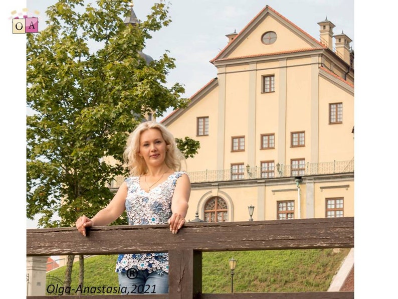 May include: A woman with blonde hair is standing on a wooden bridge in front of a large yellow building. She is wearing a white and blue crocheted top and blue jeans. The building has many windows and a small round window in the roof.
