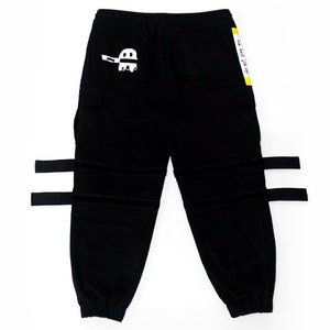May include: Black cargo pants with adjustable straps and a white graphic on the left leg. The graphic is a ghost with a striped pattern. The text "One Hell Of A Life" is printed on a yellow tag attached to the right side of the pants.