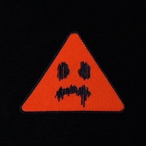 May include: A red triangle warning sign with a black embroidered sad face.