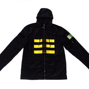 Puede incluir: Chaqueta negra con capucha, cierre de cremallera y correas amarillas en el pecho. La chaqueta tiene un pequeño parche en la manga con un diseño azul, amarillo y verde.