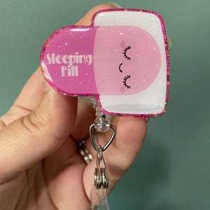 May include: Pink glitter badge holder shaped like a sleeping pill with the text "Sleeping Pill" and a cartoon face with eyelashes.