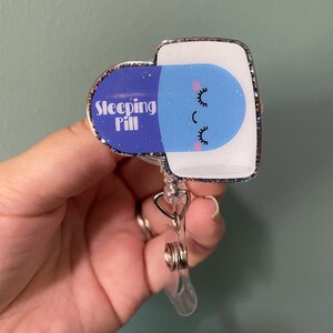 May include: A badge reel with a glittery border featuring a blue and white sleeping pill design. The pill has a smiling face with closed eyes and the words "Sleeping Pill" printed on it. The reel has a clear plastic clip and a metal retractable cord.
